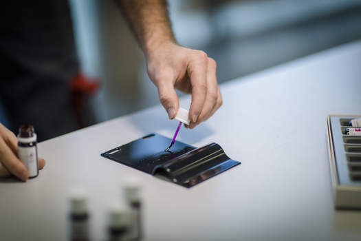 Person trägt mit Pinsel eine Flüssigkeit auf eine lackierte Probeplatte auf.JPG